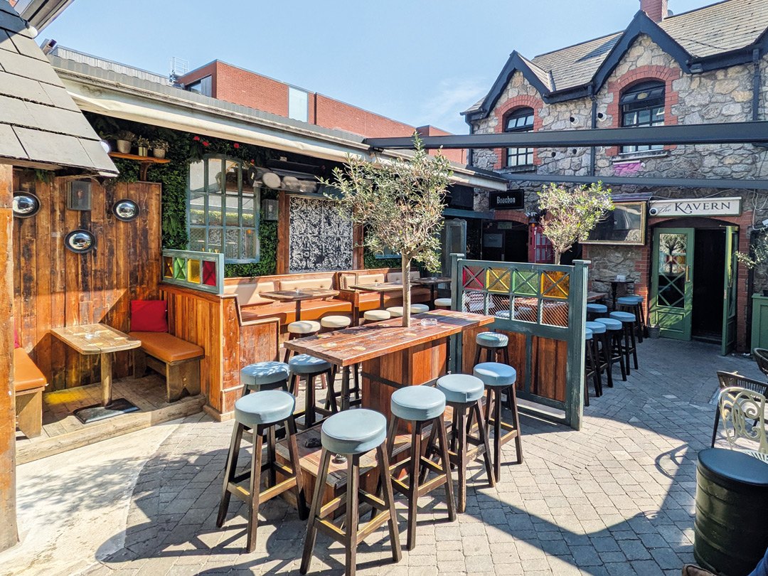 The beer garden with wooden tables, stools, plants and the stone building behind Kavanaghs