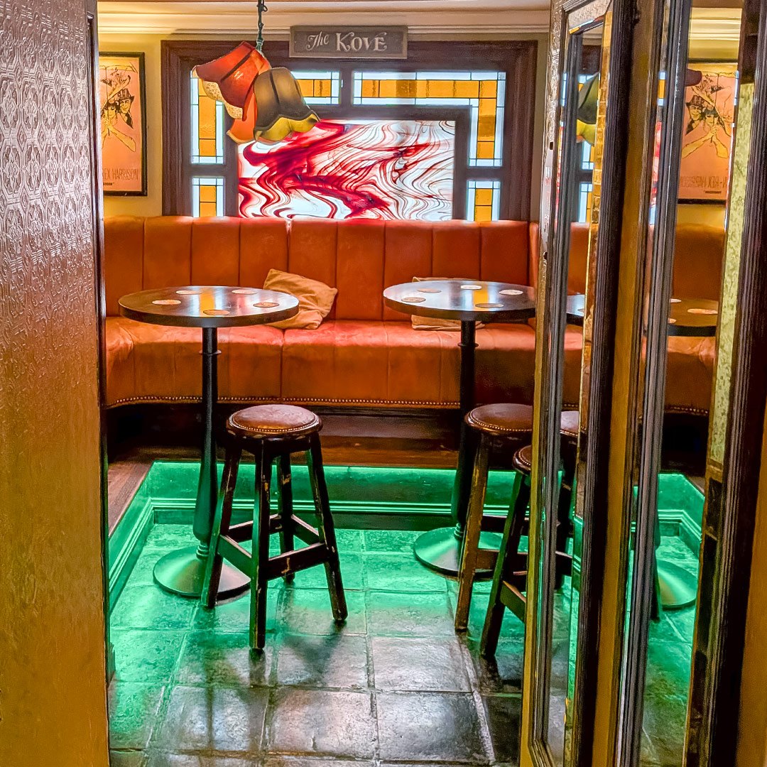 A cosy corner in The Kove with orange upholstered seats, round tables and a stained-glass window